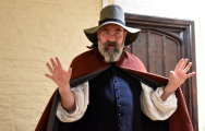Man in medieval outfit with large hat and cape explaining something